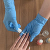 A hand wearing a blue nitrile glove is using a white applicator to apply product onto another person's fingers.