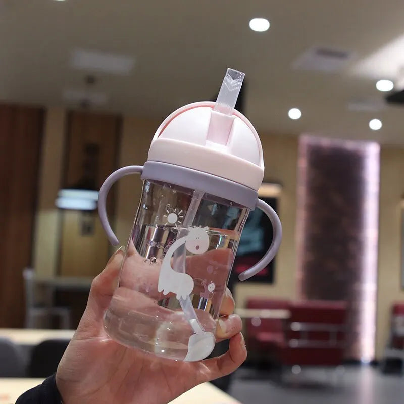 A hand holds a clear plastic sippy cup with a pink lid and gravity straw against a blurred indoor background.