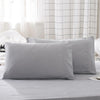 A set of two light gray microfiber pillowcases resting on a bed with white curtains in the background.