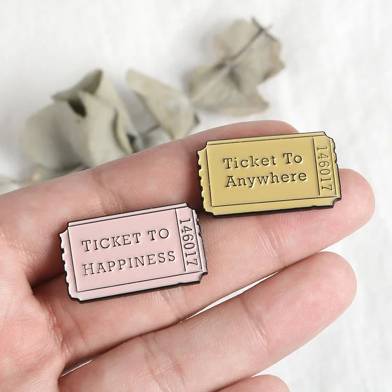 A hand holds two vintage airline ticket enamel brooches, one labeled Ticket to Anywhere and the other Ticket to Happiness.
