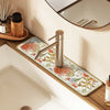 A modern metal faucet mat with a floral design is placed on a wooden bathroom counter.
