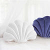 A white and purple velvet shell print throw pillow resting on a white bed with sheer curtains.