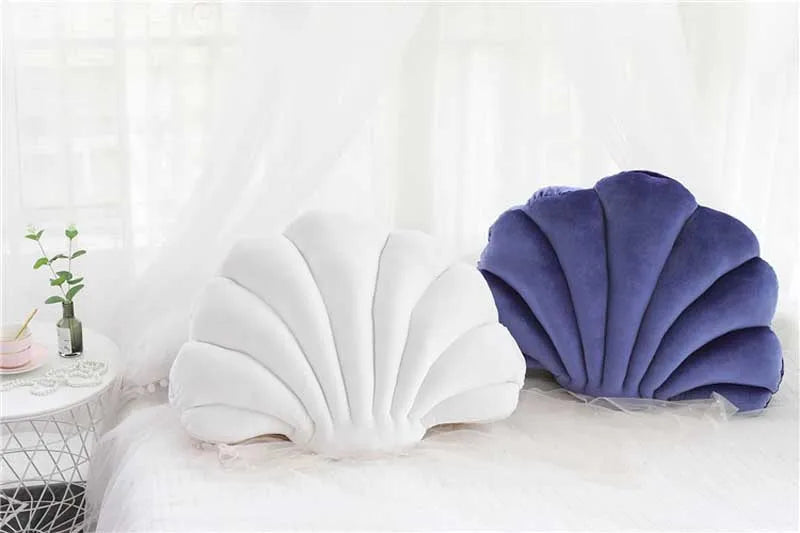 A white and purple velvet shell print throw pillow resting on a white bed with sheer curtains.