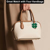 Helles beige faltbares Handtasche mit braunem Lederriemen und grünem Kleeblatt-Design, gehalten von einer Hand.