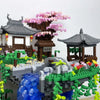 A miniature building block scene featuring a traditional Japanese-style house, a pink sakura tree, and a flowing blue water f