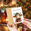 A set of Christmas cards featuring an adorable Highland cow design, held by a person in a festive indoor setting.