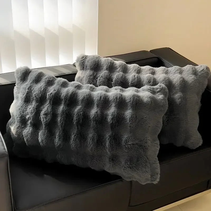A gray, fluffy quilted velvet pillow is displayed on a black sofa.