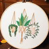 Floral cross stitch embroidery kit featuring a potted plant with green leaves and a wreath, displayed on a white canvas.