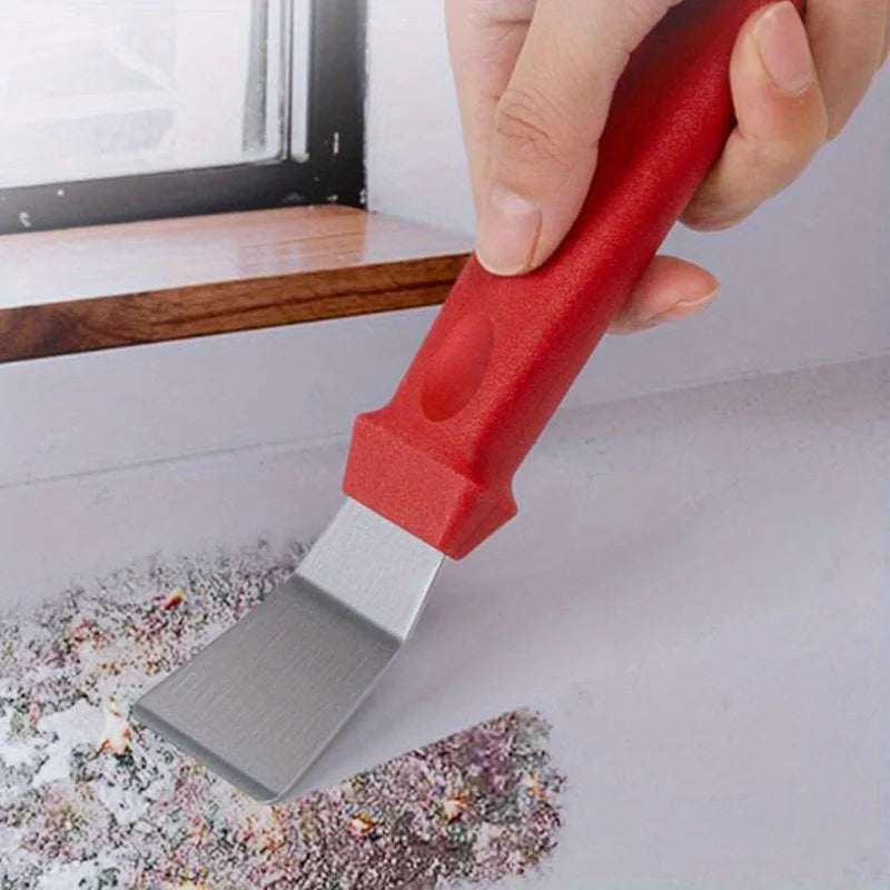 A hand holds a red kitchen cleaning brush set with a metal scraper tool resting on a light surface.