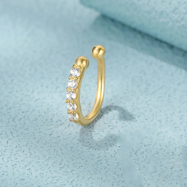 A gold-colored geometric nose cuff with small clear stones is shown against a light blue background.