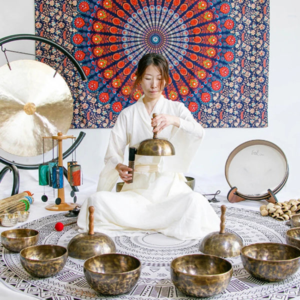 A person sits in a traditional style, surrounded by handcrafted Tibetan singing bowls and a wooden mallet against a colorful 