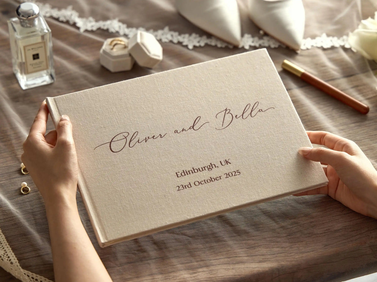 A close-up of a personalized linen wedding guest book with elegant script, sitting on a wooden surface next to a perfume bott