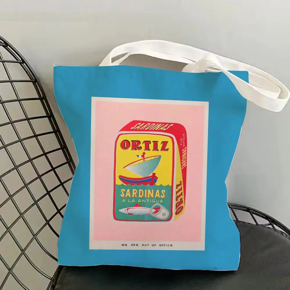 A blue canvas tote bag with a pink label featuring a graphic of sardines and the text Ortiz Sardinas a la Antigua