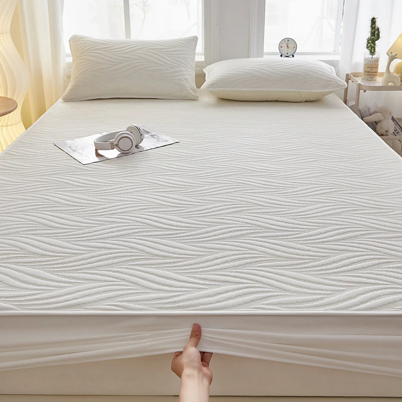 A white fitted mattress protector with a textured, wavy pattern is shown on a bed, next to pillows and a clock.