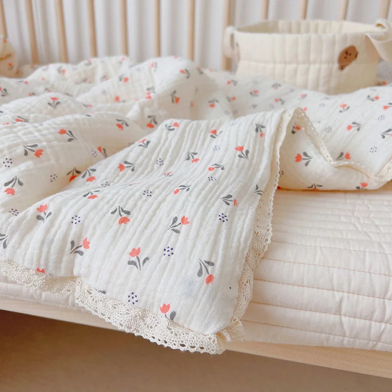 A white floral muslin baby blanket with small orange and gray flowers is folded on a light pink surface.