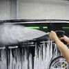A hand is using a heavy-duty pressure washer gun with a foam cannon to spray black and white paint on a vehicle's surface.