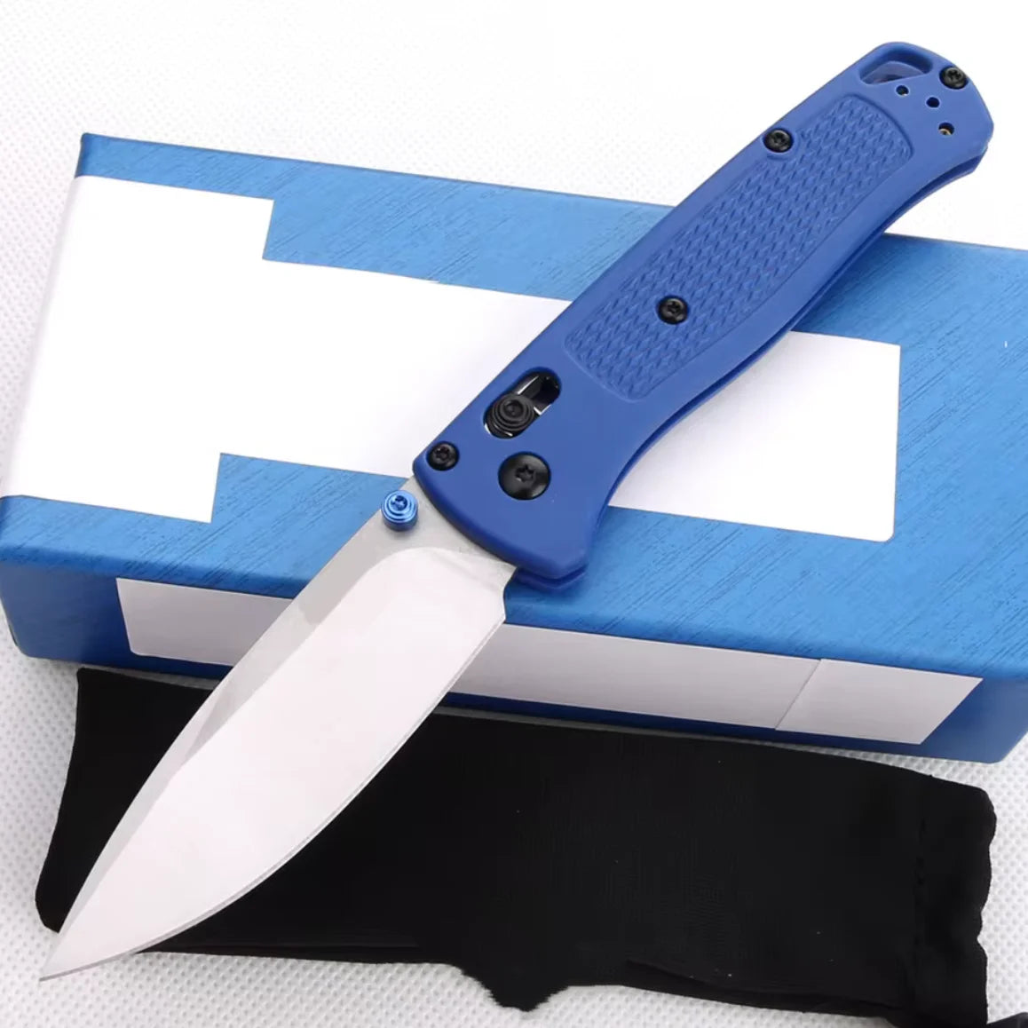 A blue compact folding knife with a stainless steel blade is shown resting on a white box and a black sheath.