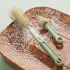 A silicone baby bottle brush set with a light green handle and bristles is shown on a wooden board surrounded by small white 