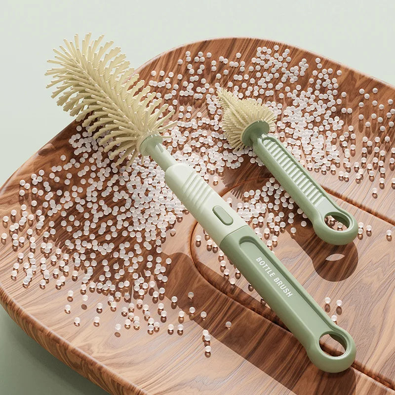 A silicone baby bottle brush set with a light green handle and bristles is shown on a wooden board surrounded by small white 