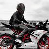 A person wearing a black full face motorcycle helmet is sitting on a black and white sport bike against an overcast sky.