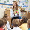 A smiling young woman sits in front of a bulletin board displaying a wooden clock toy for children.