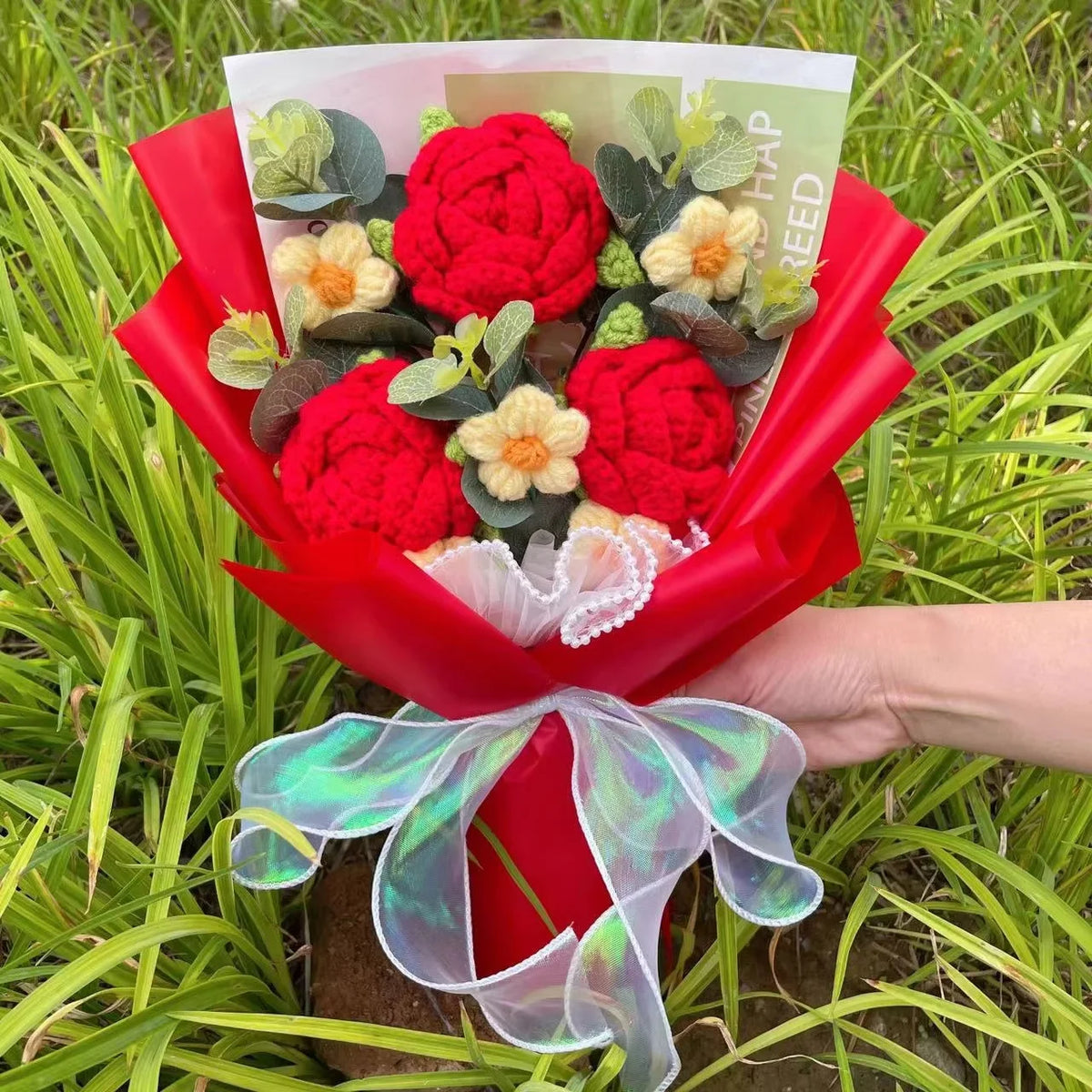 Ein Strauß roter Häkelblumen und grüner Belaubung, in rohem Papier eingewickelt und mit einem durchsichtigen grünen Band gebu