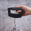 A hand holds a black portable digital luggage scale with a backlit display showing Max: 50kg d=10g.