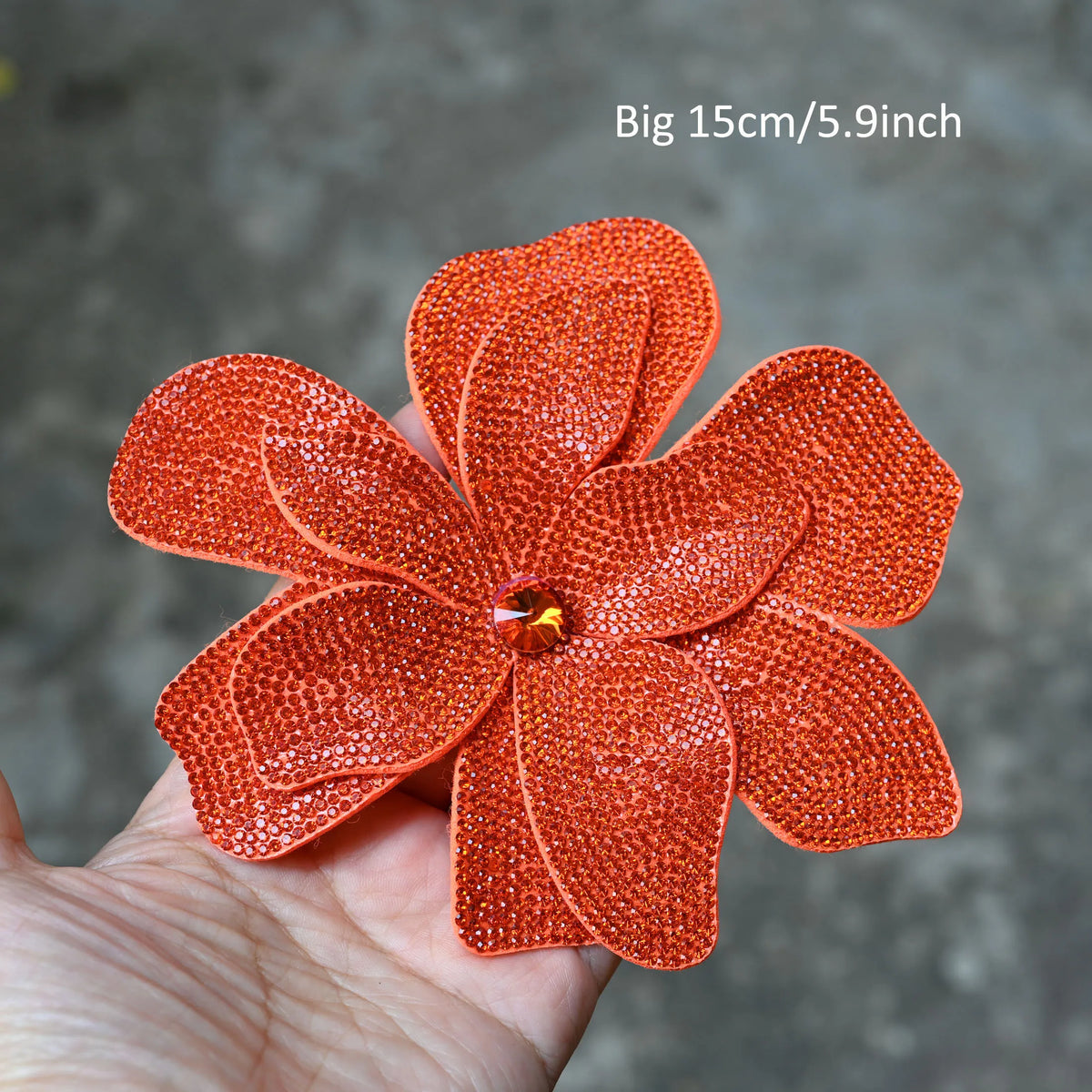 A hand holds an orange crystal rhinestone flower sew-on patch for clothing and accessories.