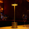 A modern gold-toned desk lamp with an illuminated circular light, sitting on a wooden surface next to a glass of red wine.