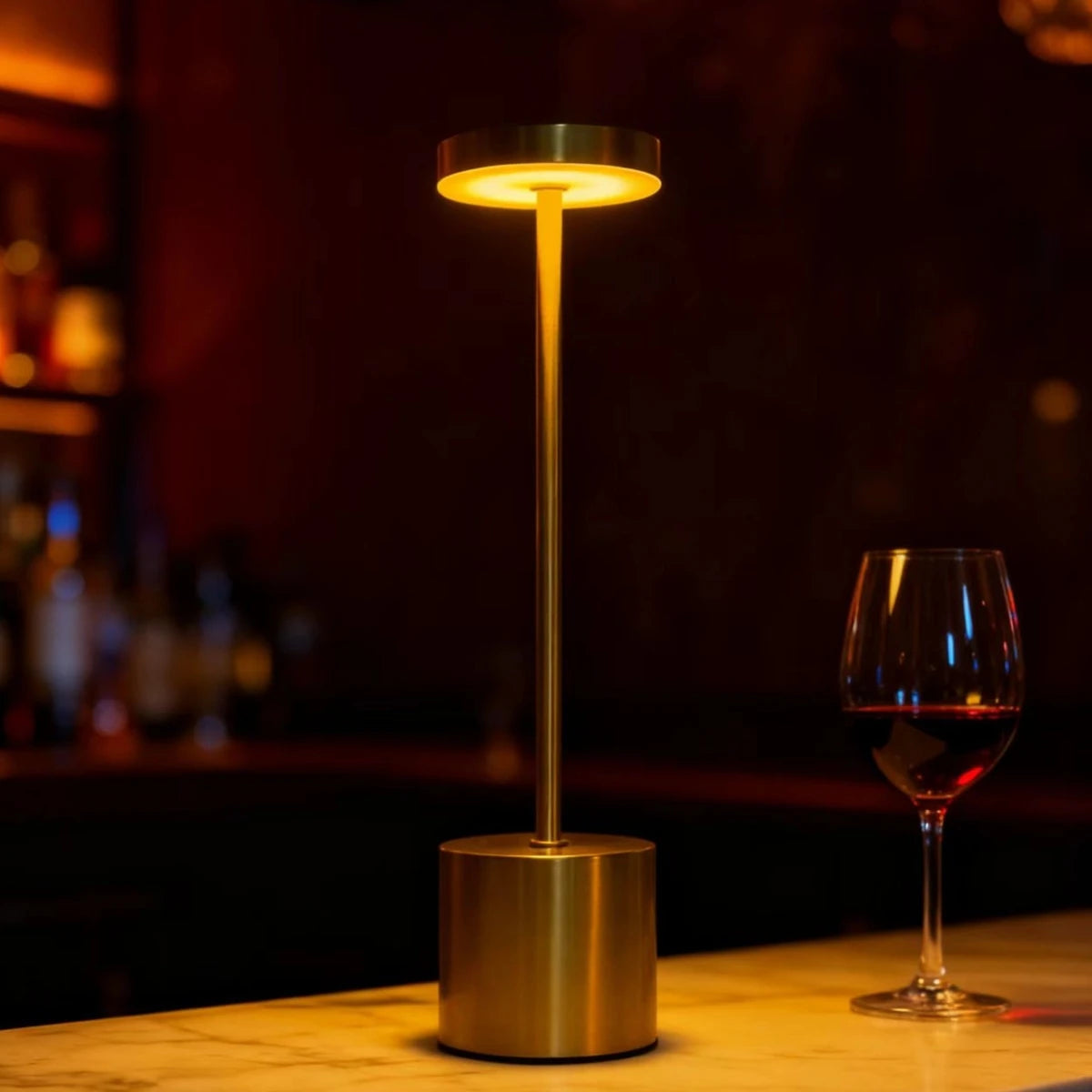 A modern gold-toned desk lamp with an illuminated circular light, sitting on a wooden surface next to a glass of red wine.