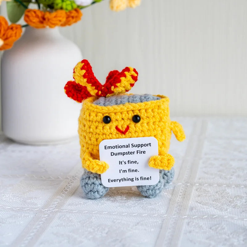 Handmade crochet doll resembling a dumpster fire with bright yellow body and red hair, holding an emotional support sign.
