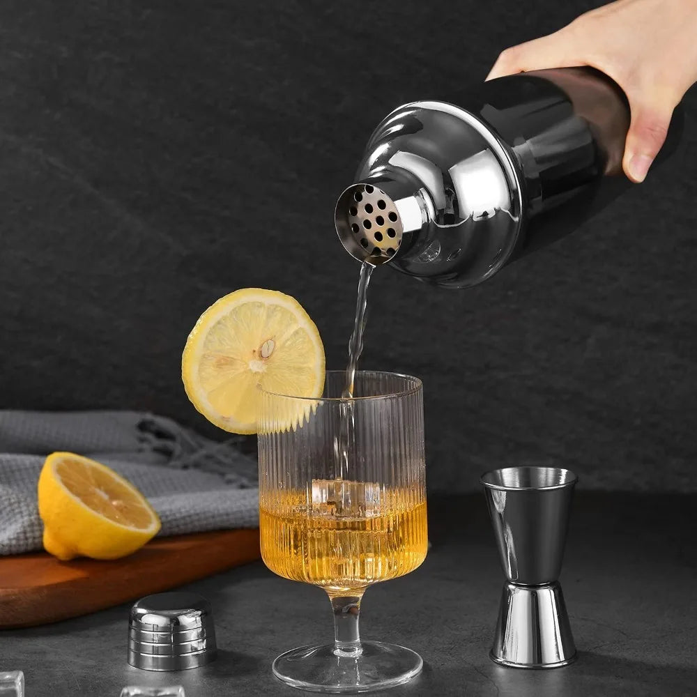 A hand pours a slice of lemon into a glass of amber-colored cocktail from a stainless steel shaker.