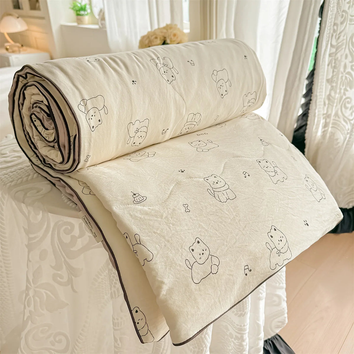 A cream-colored quilt with a pastoral print featuring small animal figures rolled up on a white surface.
