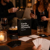 A close-up of a table with cards and glasses, showing people interacting with the Absurd Box party game.