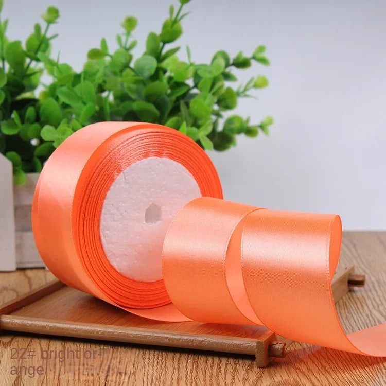 A roll of bright orange satin ribbon lying on a wooden surface with green foliage in the background.