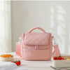 A pink quilted insulated lunch bag is displayed on a white table next to some fresh fruit and an open book.