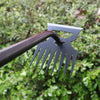 A close-up of a black stainless steel weeding shovel and rake being used in green foliage.
