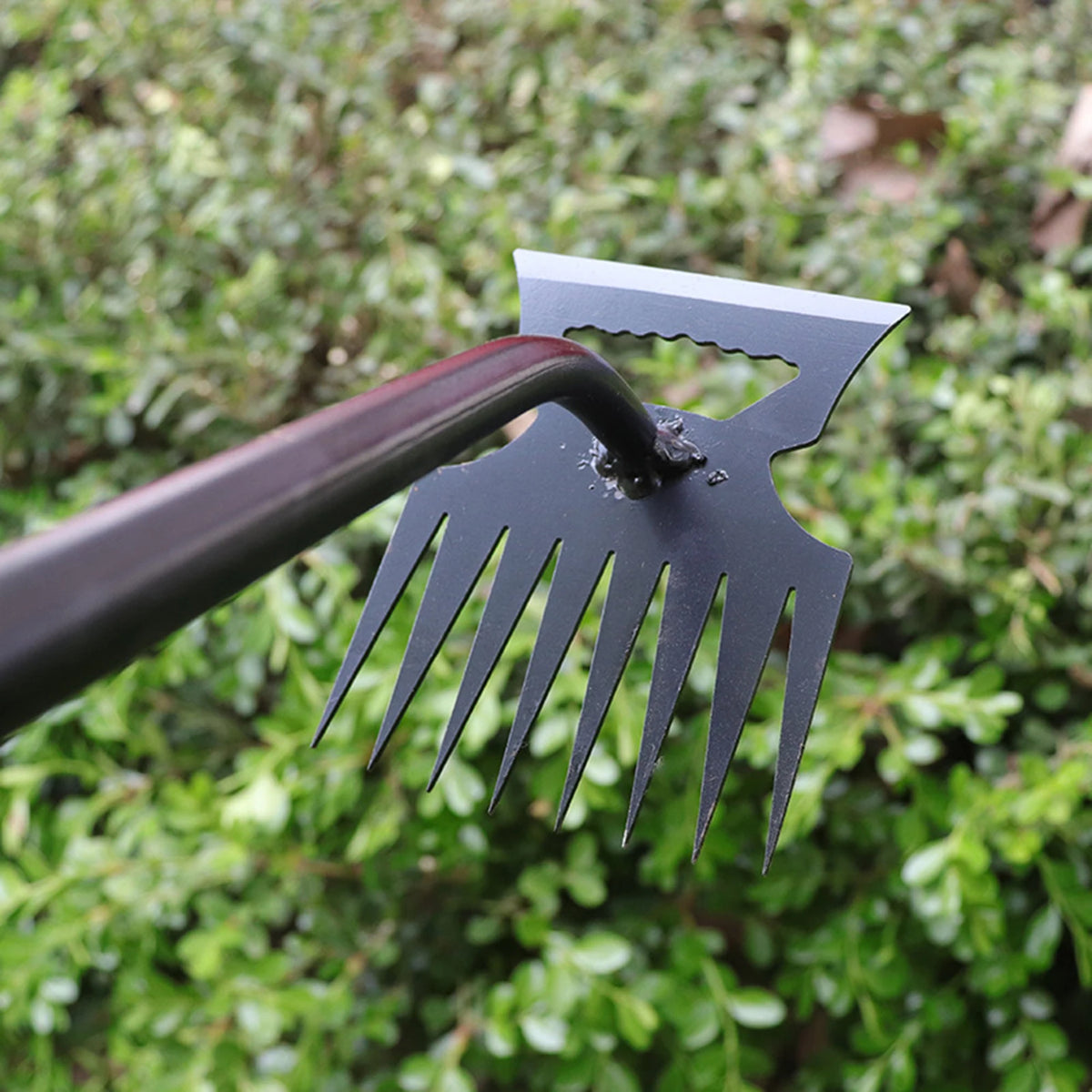 A close-up of a black stainless steel weeding shovel and rake being used in green foliage.