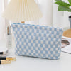 A light blue and white checkered plaid makeup bag with a zipper lying on a white surface next to a lamp and cosmetic products