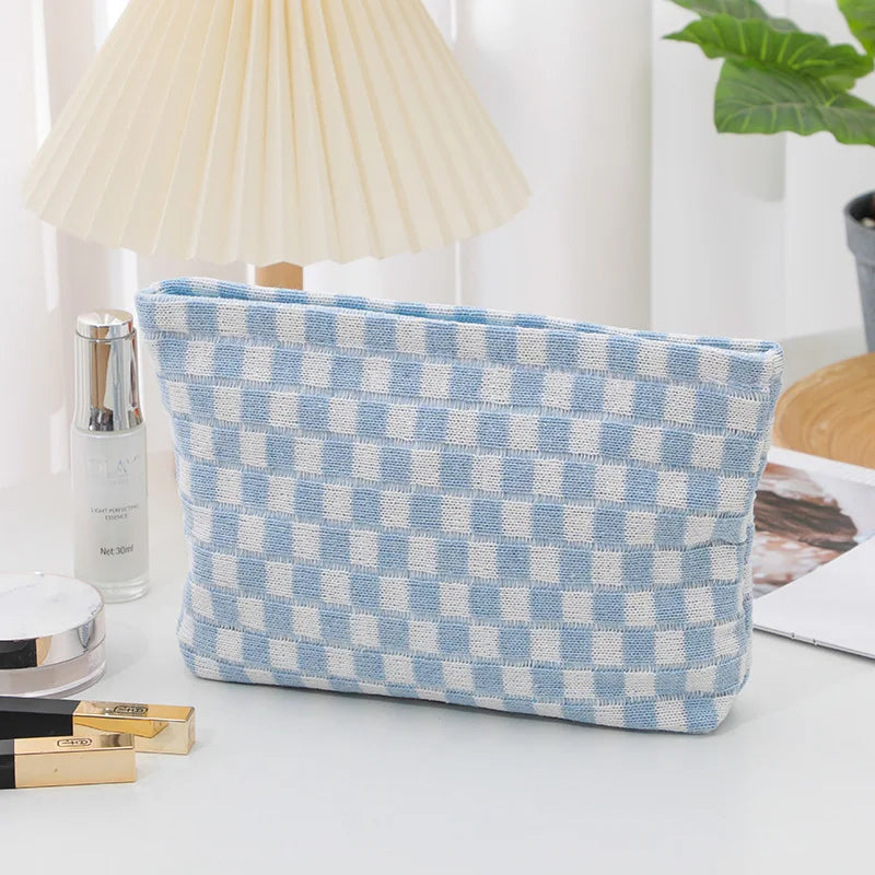A light blue and white checkered plaid makeup bag with a zipper lying on a white surface next to a lamp and cosmetic products
