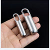 A hand holds a stainless steel waterproof pill capsule with a keychain attachment.