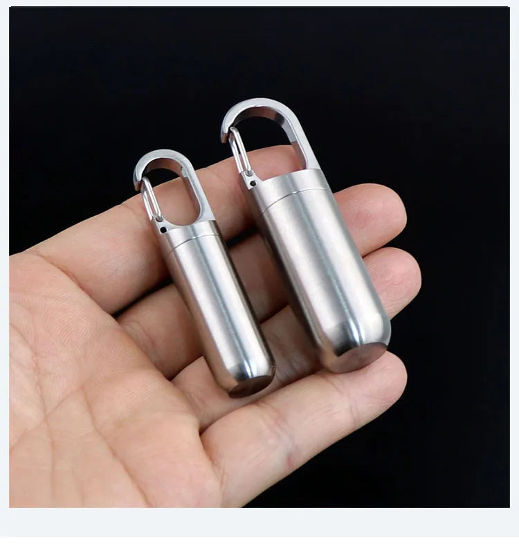 A hand holds a stainless steel waterproof pill capsule with a keychain attachment.