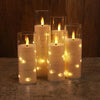 A set of glowing white candles in glass holders decorated with twinkling string lights and a visible flame.