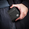A hand is holding a black RFID-blocking travel wallet with a carabiner clip inside a dark garment pocket.