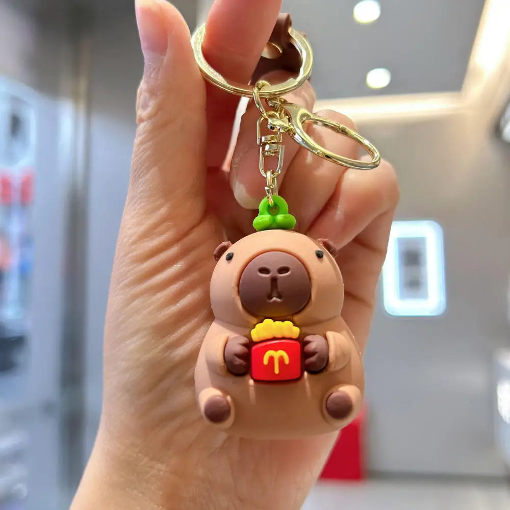 A hand holds an adorable capybara keychain charm with a green top and a red box detail.