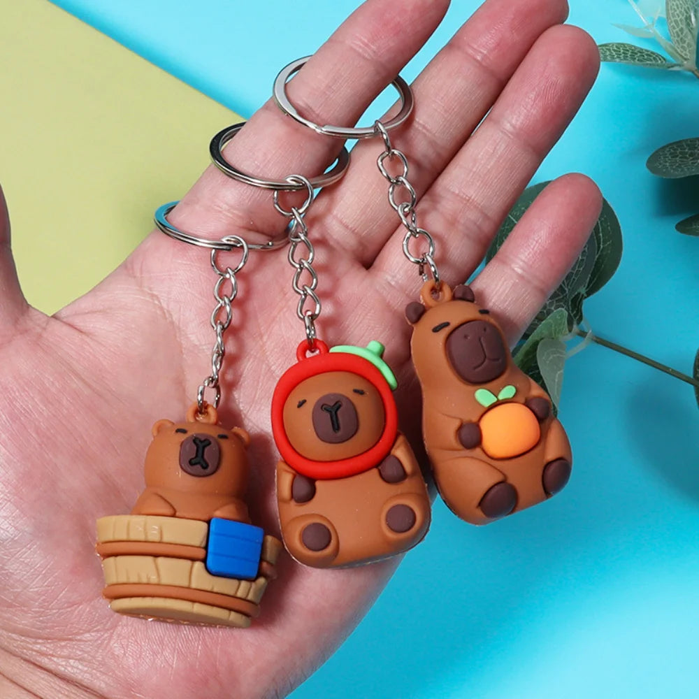 A hand holds an adorable capybara keychain charm with a wooden body and orange ear, attached to a silver ring.