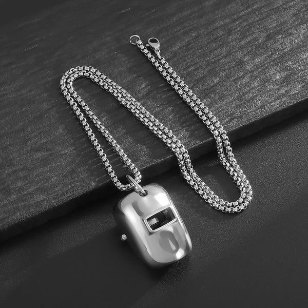 Silver stainless steel welder mask pendant necklace with a chain and bail resting on a dark surface.
