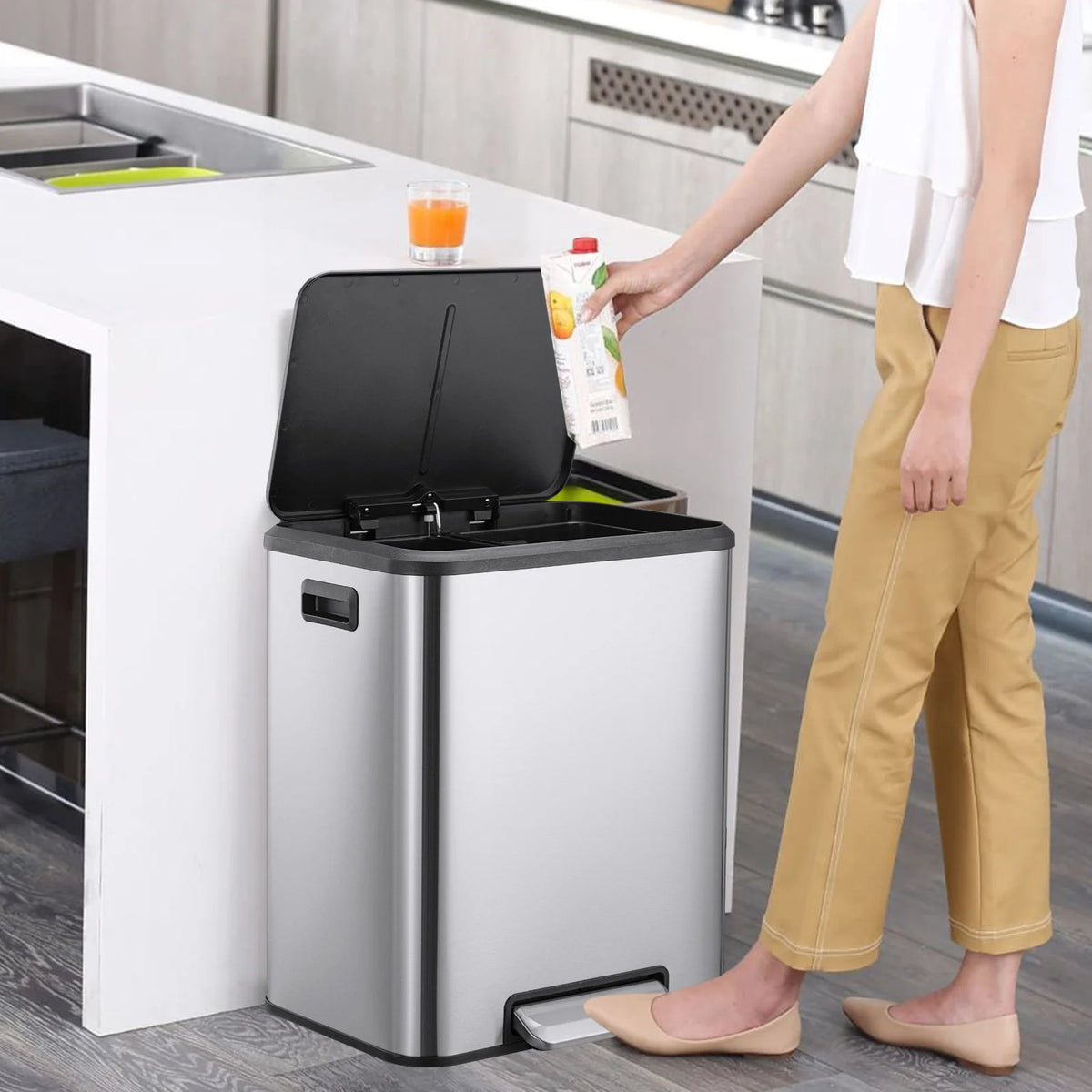 A person is opening a stainless steel dual compartment trash can with a soft-close lid in a kitchen setting.
