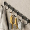 A set of six bronze wall hooks with adhesive backing are mounted on a gray wall, holding various brushes and tools.