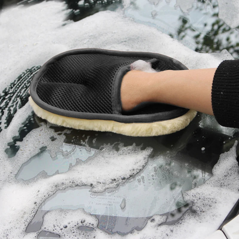 A hand wearing a black microfiber car wash glove is shown cleaning a car surface covered in foam.